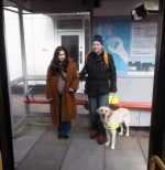 A woman guiding a man with sight loss at a bus stop, who is with their guide dog.