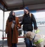 A woman guiding a man with sight loss at a bus stop, who is with his guide dog.